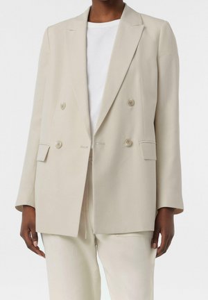 Personne portant un blazer beige clair croisé avec un pantalon assorti et une chemise blanche à col rond, visible du cou jusqu'à mi-cuisse.
