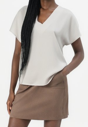 Femme portant un haut blanc ample à col en V et manches courtes, ainsi qu'une jupe marron avec des poches, posant avec une main dans une poche.