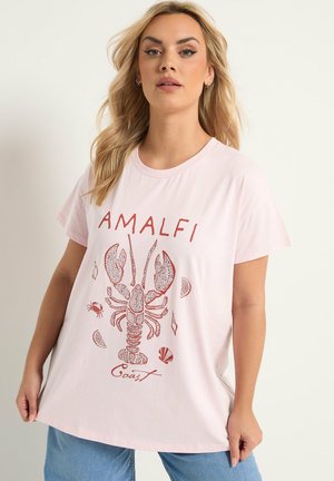 Mujer vistiendo una camiseta rosa claro con una ilustración de una langosta roja y el texto "Costa Amalfi", combinada con jeans azules, posando frente a un fondo liso.