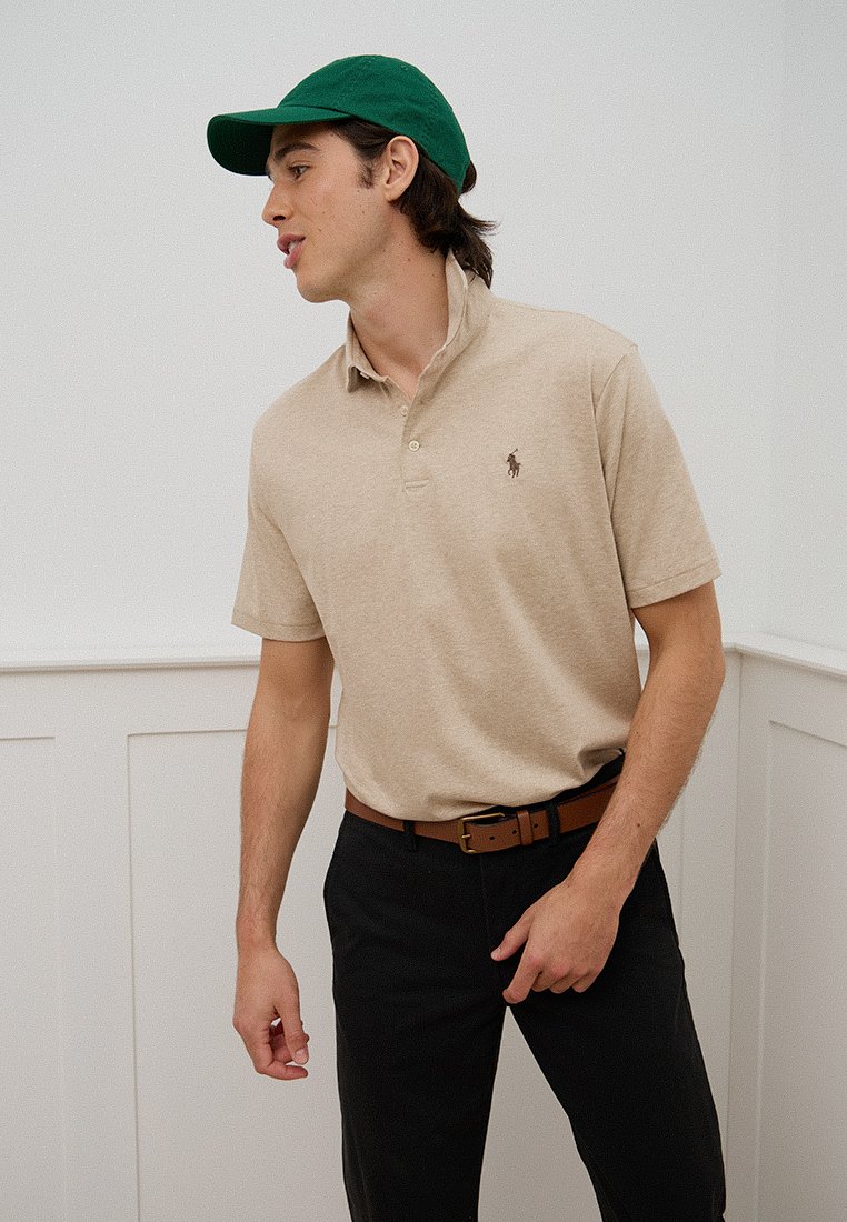 Un jeune homme en polo beige et pantalons noir, portant une casquette verte, se tenant contre un mur blanc à panneaux, regardant vers la gauche.