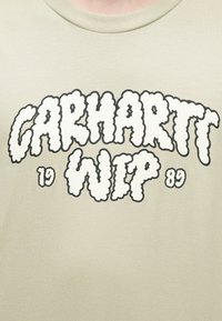 Μπεζ βαμβακερό t-shirt με έντονα, σύννεφο-σχήματος μαύρα και λευκά γράμματα που γράφουν "CARHARTT WIP 89" στο κέντρο μπροστά.