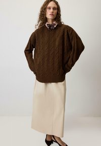 Brown cable-knit sweater with dropped shoulders, paired with a floor-length cream skirt. Model wears a patterned bowtie under the sweater.