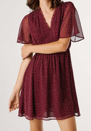 Kobieta w krótkiej bordowej sukience w białe kropki, z przezroczystymi falbankami na rękawach i dekoltem w kształcie litery V, stojąca z jedną skrzyżowaną ręką.