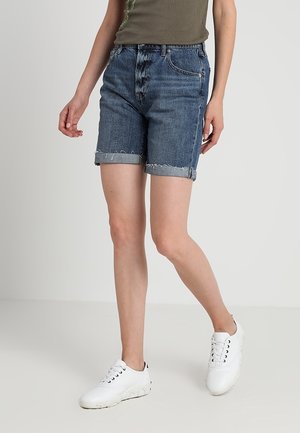 Vrouw die blauwe denim shorts met opgerolde zoom draagt, een olijfgroene mouwloze top en witte casual sneakers, staand tegen een effen lichte achtergrond.