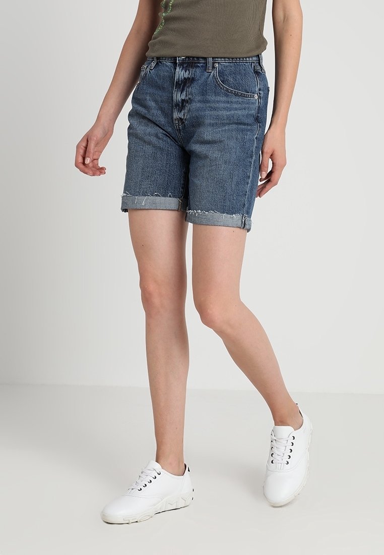 Femme portant un short en jean bleu retroussé, un haut sans manches vert olive et des baskets blanches décontractées, debout contre un fond clair uni.