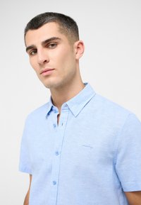 Camisa de botón de manga corta de color azul claro con un tejido texturizado, un cuello puntiagudo y un pequeño logo bordado en el lado izquierdo del pecho.