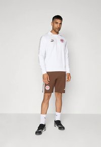 Mies seisoo valkoisessa pitkähihaisessa puolipolopaidassa, ruskeissa logollisissa shortseissa, valkoisissa sukissa ja mustissa tennareissa vaaleaa taustaa vasten.