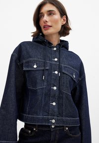 Kratka jeans jakna v temno modri barvi s kapuco, srebrnimi gumbi, dvema žepoma na prsih in kontrastnim belim šivanjem.