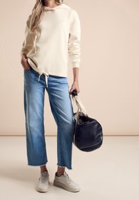 Kremowa bluza z kapturem i sznurkiem, niebieskie szerokie dżinsy oraz białe sneakersy. Modelka trzyma okrągłą, granatową torbę z jasnymi paskami.