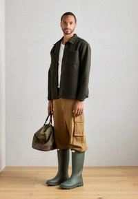 Bottes en caoutchouc vertes, pantalons cargo marron, veste à boutons olive sur une chemise blanche. Sac de sport marron et olive tenu à la main.