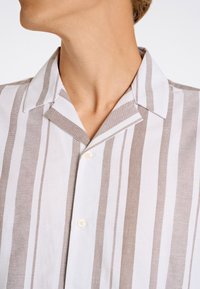 Camicia a maniche corte con bottoni a righe bianche e beige; presenta un colletto da campeggio, un tessuto leggero e una chiusura con un solo bottone.