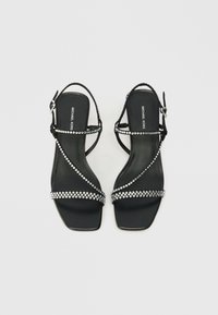 Czarne sandały na obcasie z kwadratowym noskiem, cienkimi paskami ozdobionymi cyrkoniami oraz zapięciem na kostce. Teksturowana wkładka z logo.
