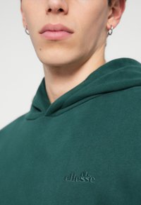 Roheline kapuutsiga sviiter, mis on valmistatud pehmest kangast, millel on rindkeres peenelt reljeefne logo ja mugav lõige.