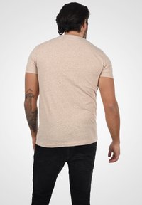 Camisa de manga corta en color beige claro con un patrón texturizado, combinada con jeans negros. Presenta un cuello redondo y un corte ajustado.