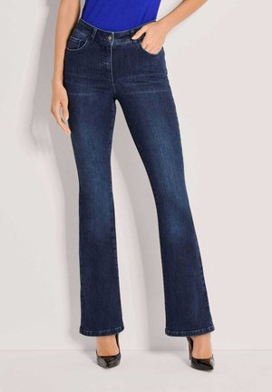 Femme portant un jean bootcut bleu foncé et des talons hauts noirs, une main dans la poche et un haut bleu partiellement visible.