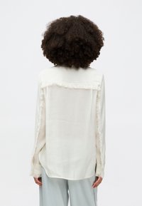 Hellescremeshirt mit Fransenbesatz an den Schultern und Ärmeln, aus einem glatten, leichten Stoff, mit relaxter Passform.