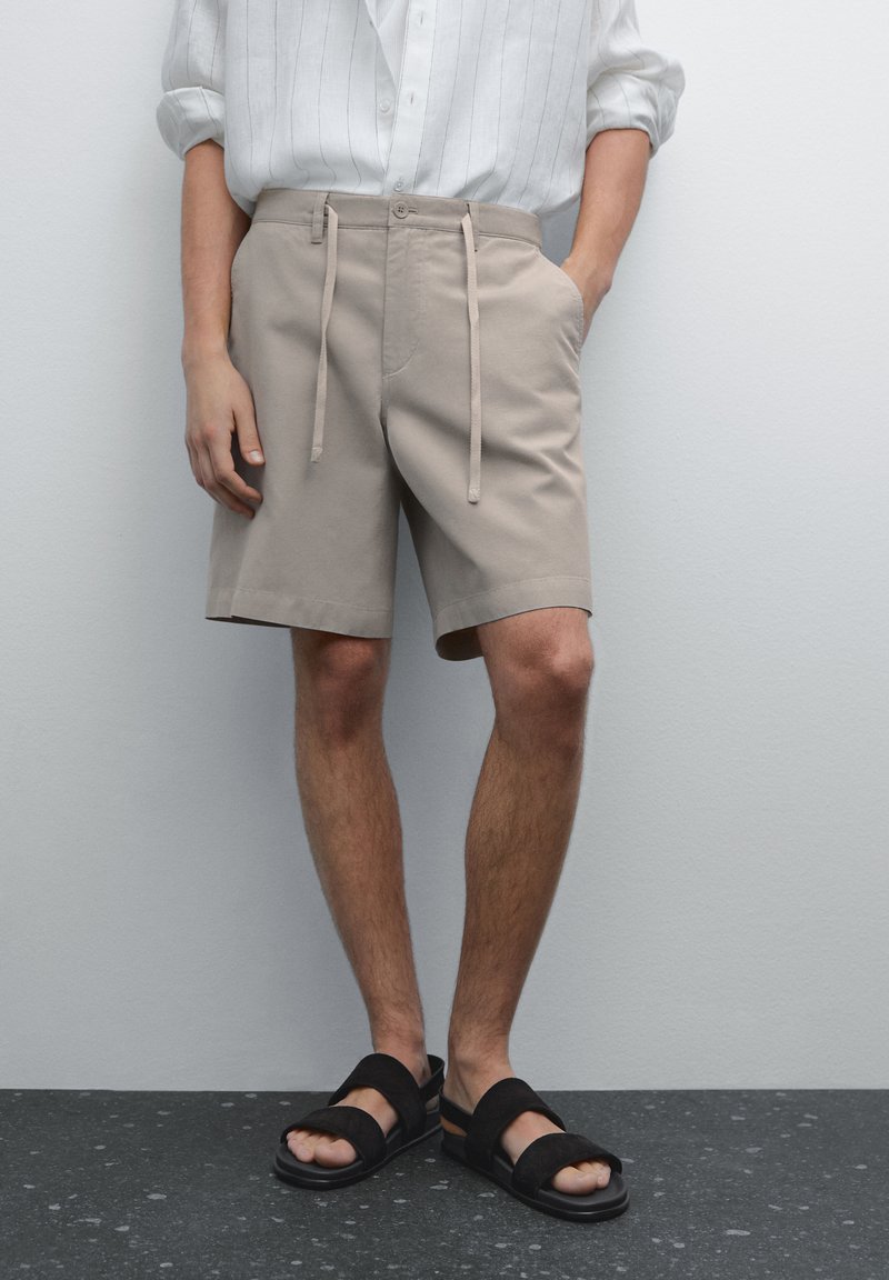Beige shorts med snoretræk i taljen, sidelommer og en afslappet pasform, parret med sorte flade sandaler, placeret mod en ensfarvet baggrund.