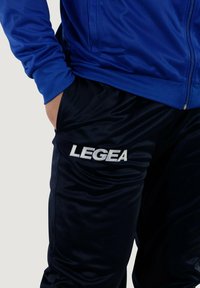 Giacca blu a zip con una texture liscia abbinata a pantaloni da jogging neri con il logo bianco "LEGEA" sulla gamba sinistra.