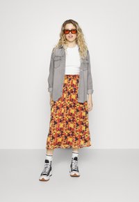 Bloemige midi rok met een rood, oranje en geel patroon, gecombineerd met een grijs button-up shirt en zwarte high-top sneakers. Inclusief oranje zonnebril.