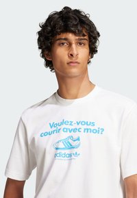 White t-shirt featuring blue text reading “Voulez-vous courir avec moi?” with an illustration of a sneaker and the Adidas logo.