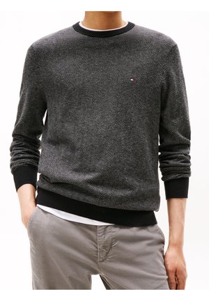 Homme portant un pull à motif chevrons gris foncé avec des poignets et un ourlet noirs, associé à un pantalon gris clair.
