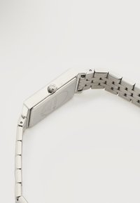 Montre rectangulaire en argent avec un bracelet à maillons métalliques, vue de côté mettant en avant sa couronne et les détails du boîtier arrière.
