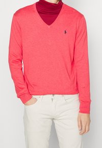 V-ringad tröja i ljus korall, stickad tyg, med långa ärmar och en liten logotyp på bröstet, kombinerad med vita byxor.