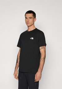 Tricou din bumbac negru, cu guler rotund, mâneci scurte și un logo alb brodat pe partea stângă a pieptului. Modelul este poziționat în profil.