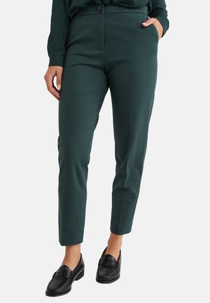 Zelené kalhoty na míru se slim fit, hladkým materiálem, bočními kapsami a knoflíkovým zapínáním v pase. Nenošeny s černými mokasínami.
