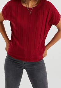 Červený ribkovaný pletený top s krátkým rukávem a kulatým výstřihem, s volným střihem a hladkým povrchem. Nosit s šedými džíny.