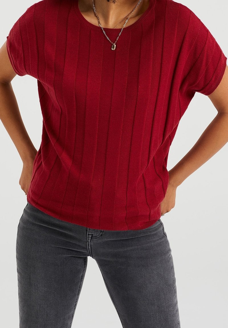 Červený ribkovaný pletený top s krátkým rukávem a kulatým výstřihem, s volným střihem a hladkým povrchem. Nosit s šedými džíny.