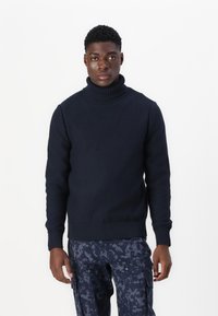 Marineblå strikket høyhalser med ribbestrikkede mansjetter og nederkant, med en teksturert finish, kombinert med mønstrede cargo-bukser.