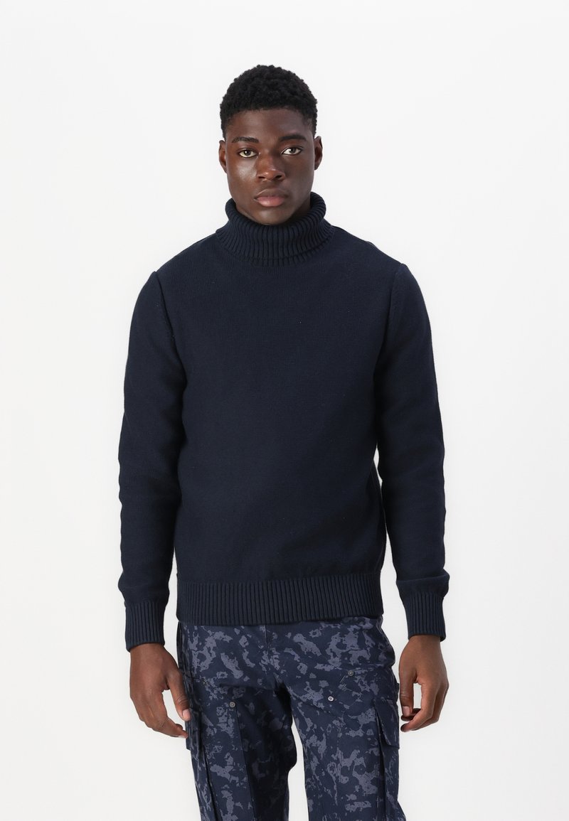 Marineblå strikket høyhalser med ribbestrikkede mansjetter og nederkant, med en teksturert finish, kombinert med mønstrede cargo-bukser.