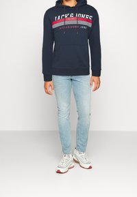 Persona vestida con sudadera azul marino con texto en rojo y blanco, jeans ajustados de color azul claro y zapatillas blancas con acentos rojos, de pie sobre un fondo claro.