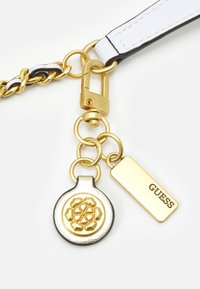 Charm de metal dorado con un diseño floral, adjunto a una correa en blanco y negro con un clip dorado. Presenta una etiqueta rectangular grabada con "GUESS."