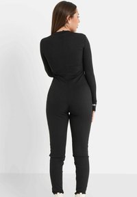 Schwarzer langärmeliger Jumpsuit mit figurbetontem Design und elastischem Stoff, mit einer Rückennaht und Logo-Print an den Ärmelabschlüssen.