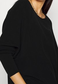 Schwarzer Strickpullover mit lockerer Passform, weitem Halsauschnitt und aufgerollten Ärmeln. Verfügt über einen strukturierten Stoff und eine goldene Kettenhalskette als Akzent.