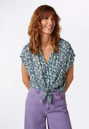 Blouse à manches courtes avec un motif floral bleu et vert, dotée d'un col en V et d'un détail noué à la taille, associée à un pantalon violet clair.