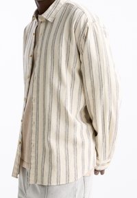 Chemise à manches longues en tissu beige avec des rayures verticales noires, dotée d'une fermeture à boutons sur le devant et d'un matériau légèrement texturé.