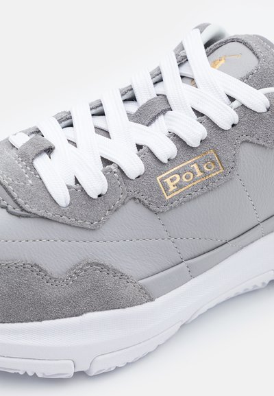 Tenis gri din piele și suede, cu șireturi albe, logo „Polo” aurie și accente texturate. Formă rotunjită a vârfului și talpă din cauciuc.