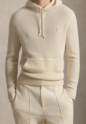 Homme portant un sweat à capuche en maille gaufrée couleur crème avec poche avant et pantalon crème sur mesure, mains dans les poches, fond neutre.