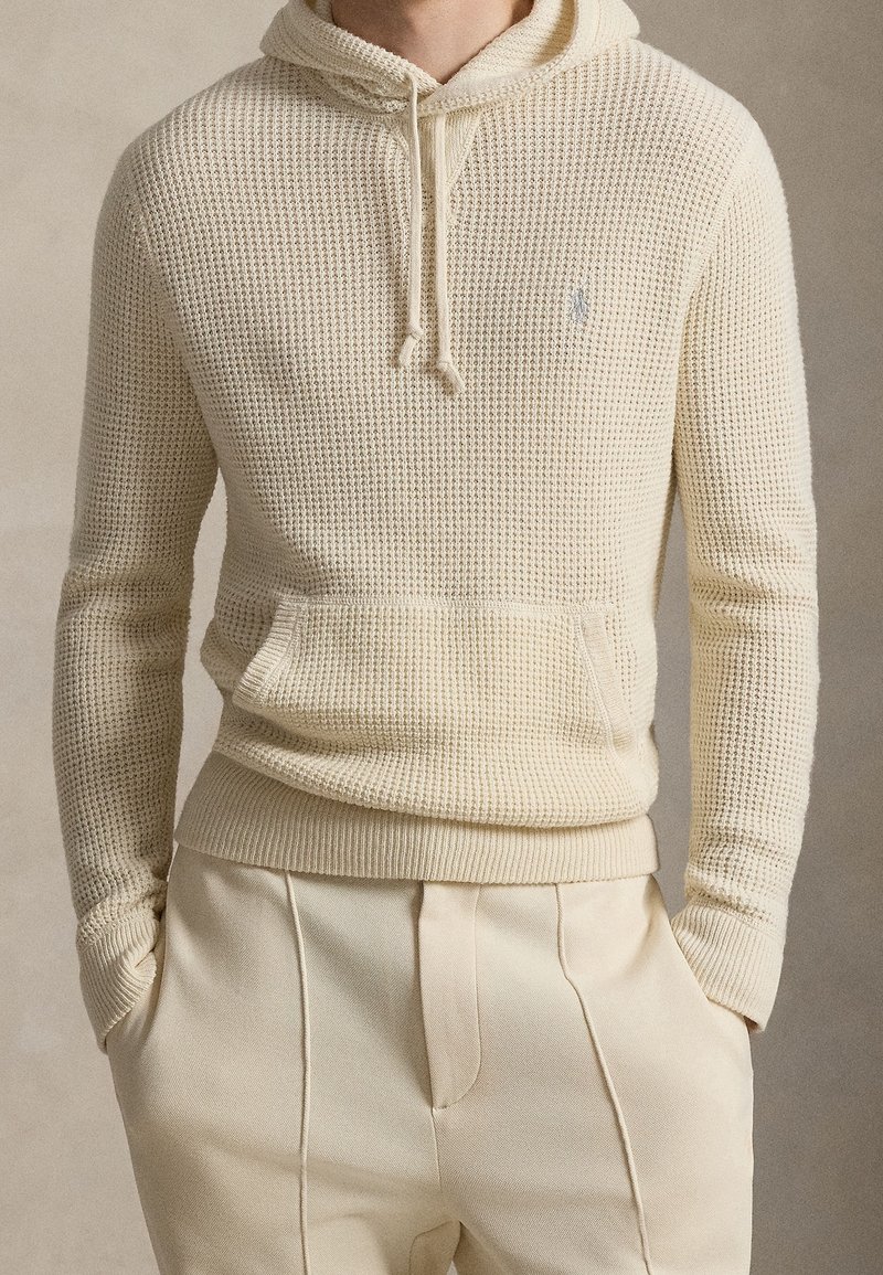 Homme portant un sweat à capuche en maille gaufrée couleur crème avec poche avant et pantalon crème sur mesure, mains dans les poches, fond neutre.