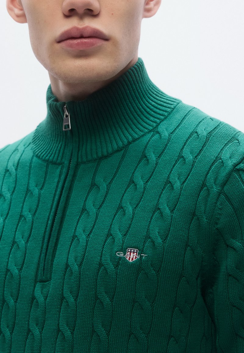 Green knit sweater with a high zip collar, featuring a cable pattern and a small logo patch on the chest.