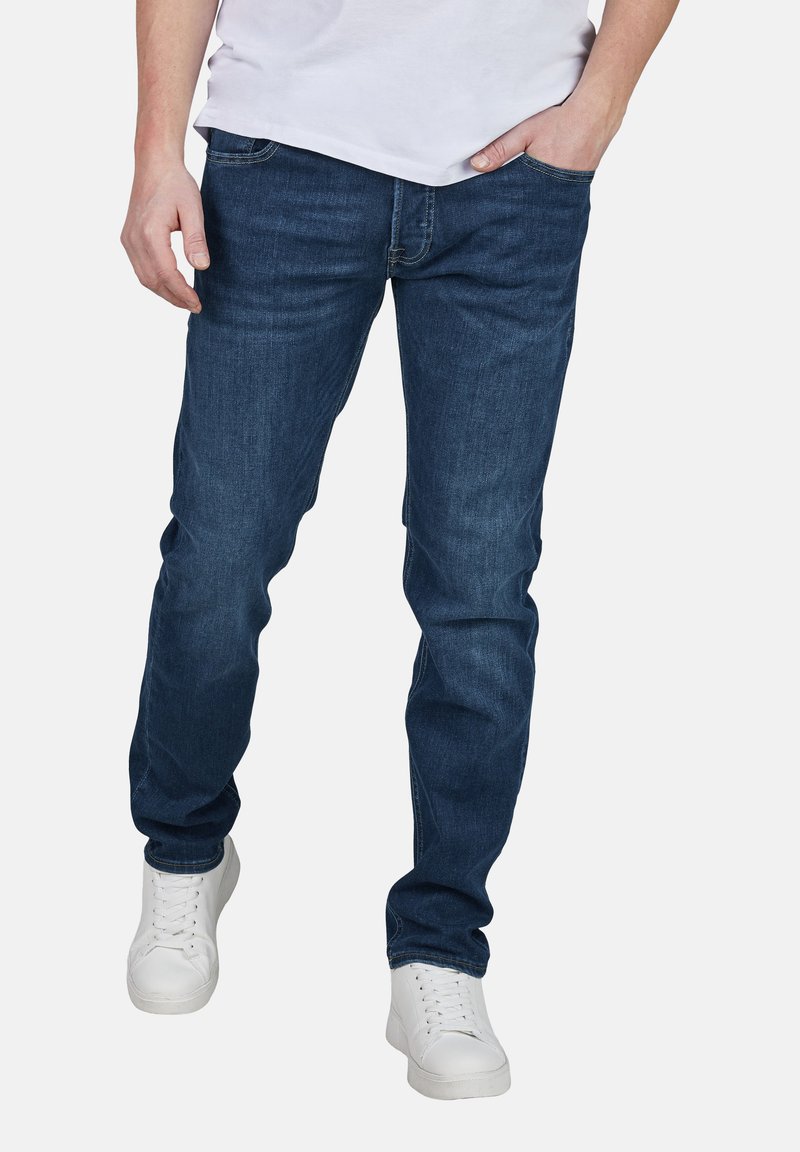 Jeans de mezclilla azul oscuro con un ajuste Slim, cinco bolsillos y un diseño clásico. Combinados con zapatillas blancas para un look casual.