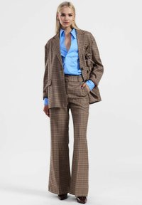 Donna che indossa un completo a quadri marroni con pantaloni svasati e un blazer oversize sopra una camicia azzurro chiaro, in piedi su uno sfondo bianco semplice.