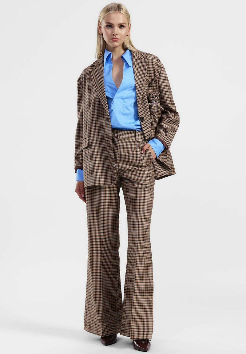 Donna che indossa un completo a quadri marroni con pantaloni svasati e un blazer oversize sopra una camicia azzurro chiaro, in piedi su uno sfondo bianco semplice.