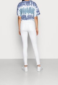 Mujer vestida con una camiseta de tie-dye azul y blanca, jeans ajustados blancos y zapatillas de plataforma blancas, de pie frente a una pared simple.