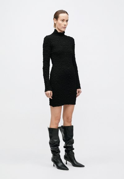 Naine seisab valgel taustal musta tekstuurse pika varrukaga minikleidi ja mustade teravate ninaga lahtiste põlvikudeni saapadega.