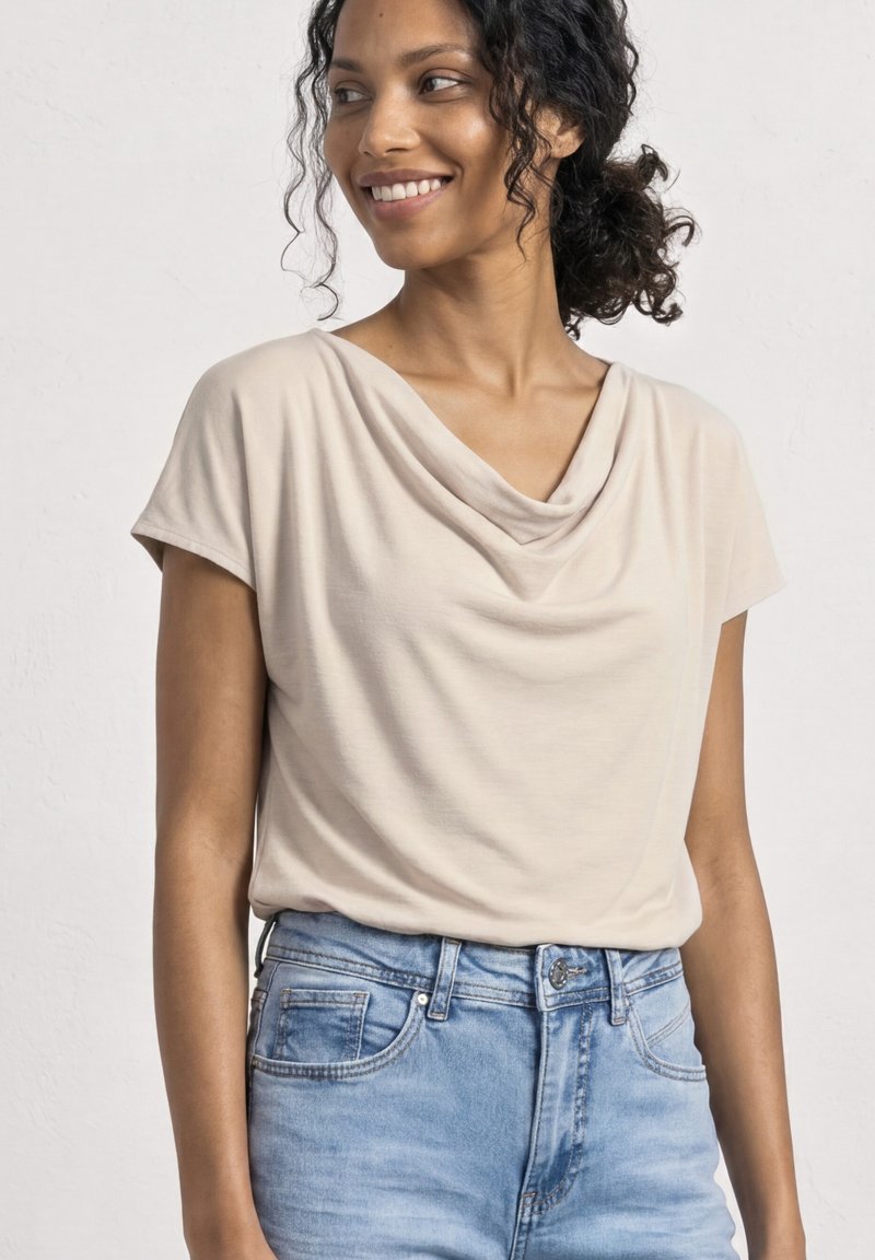 Frau mit lockigem zurückgebundenem Haar, die ein beiges Top mit Drapé-Hals und hellblaue Jeans trägt, lächelt und schaut zur Seite.