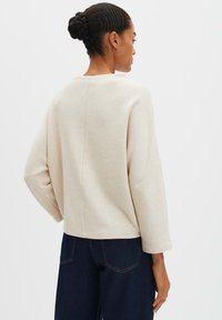 Frau mit geflochtenem Dutt-Frisur, die ein lockeres, langärmliges, beiges Oberteil und dunkelblaue Jeans trägt, steht mit dem Rücken zur Kamera vor einem weißen Hintergrund.